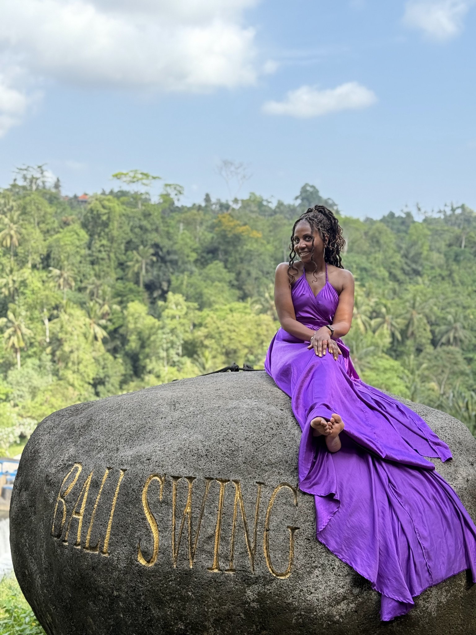 A traveler in a flowing purple gown relaxing on the iconic Bali Swing stone above the lush Ubud jungle